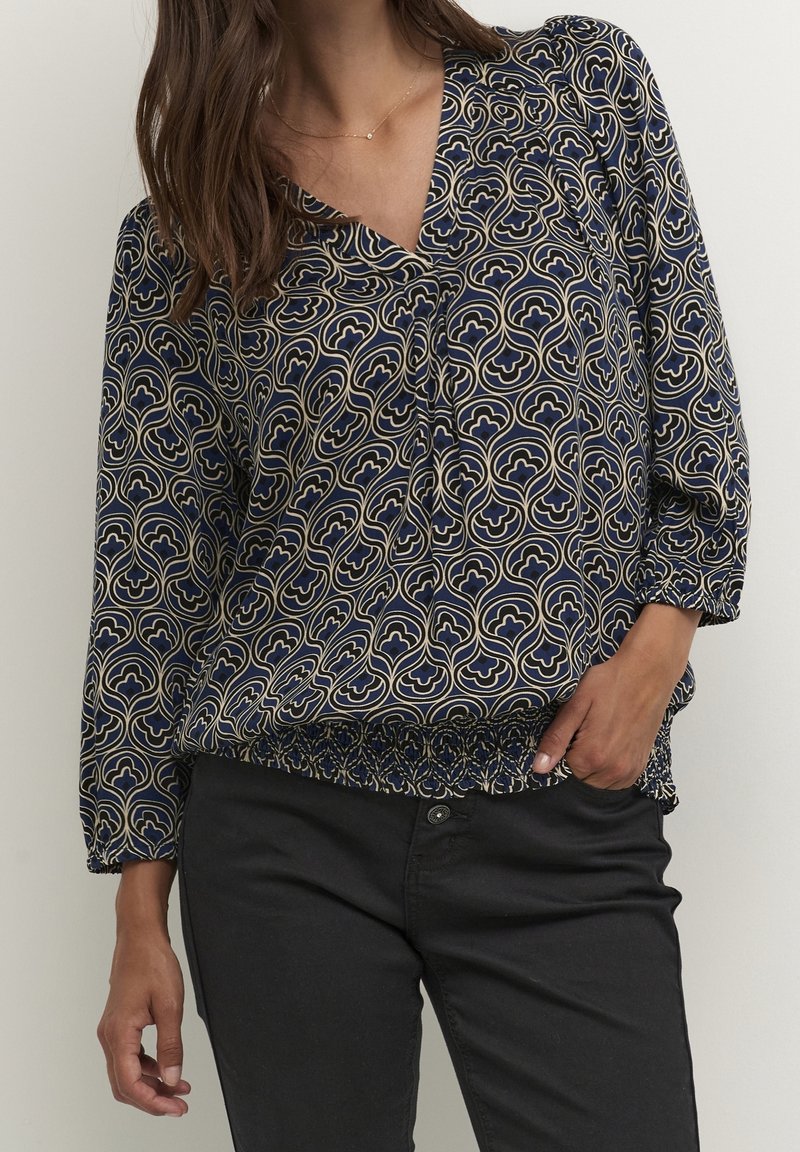Femme portant un chemisier bleu marine à manches longues avec des motifs géométriques crème et un pantalon noir, une main dans la poche, debout devant un fond uni.