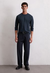 Jonge man staat tegen een witte textuurmuur, gekleed in een donker shirt met lange mouwen, een zwarte riem, donkere broek en zwarte schoenen, met het gezicht naar voren.