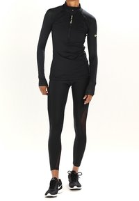 Haut de sport noir à manches longues avec col zippé, associé à un legging noir avec des panneaux latéraux transparents. Les deux articles ont une coupe ajustée.