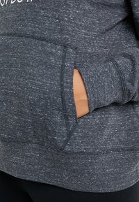 Grå huvtröja med en texturerad tyg, med en framficka och förstärkt sömnad. Designen inkluderar en logotyp på bröstområdet.