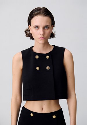 Cropped top nero realizzato in tessuto strutturato, con bottoni floreali dorati, scollatura rotonda e senza maniche. Texture liscia con design minimale.