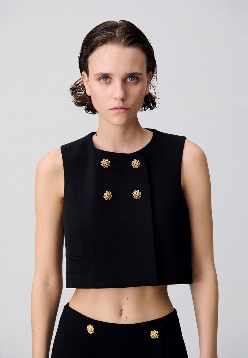 Veste noire courte en tissu structuré, ornée de boutons floraux dorés, avec un col rond et sans manches. Texture lisse avec un design minimaliste.