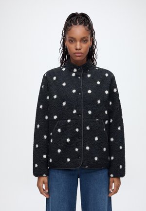 Femme aux cheveux tressés portant une veste polaire noire à pois blancs et un jean bleu, debout devant un fond clair uni.