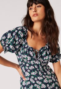 Marineblaues Blumenkleid mit Puffärmeln, das weiße und rosa Blumen auf einem grünen Akzenthintergrund zeigt, einem Rundhalsausschnitt und einer vorderen Krawatten-Details.