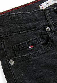 Svarta denimjeans med en liten framficka, silverdetaljer och en midjebandsetikett med röd accent. Texturerad tyg med typisk sömnad.