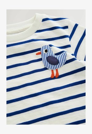 Bavlněná košile s bílými a tmavě modrými vodorovnými pruhy. Obsahuje aplikaci modrého pruhovaného ptáka s oranžovým zobákem a nohama.