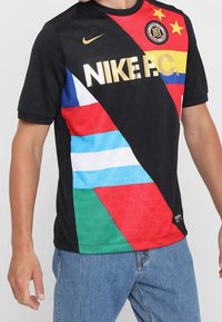 Černé tričko Nike FC s krátkým rukávem, které má diagonální patchworkový design v červené, modré, zelené a žluté barvě s zlatými akcenty loga.