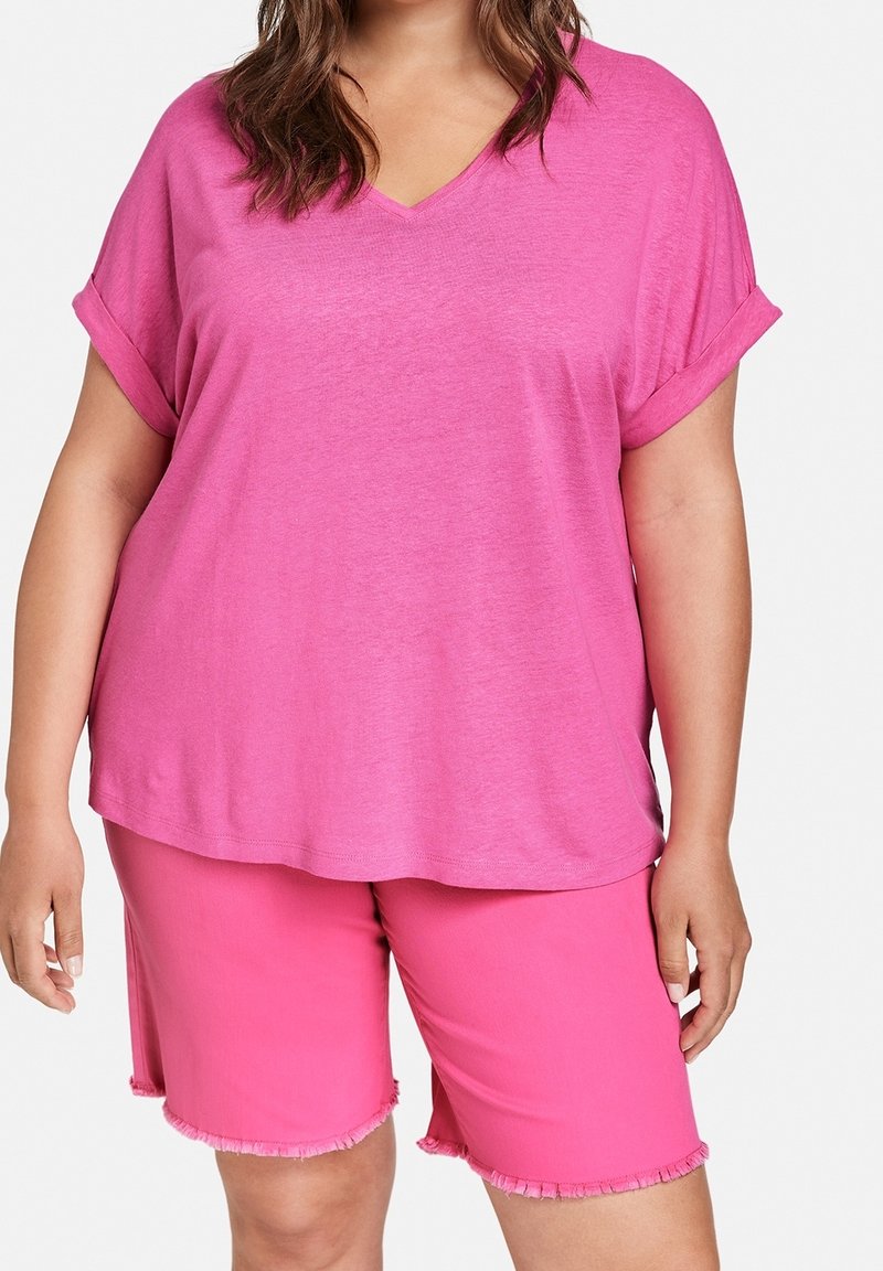 Femme porte un haut ample rose vif à manches courtes avec col en V et revers roulés, ainsi qu’un short assorti rose jusqu’aux genoux avec des ourlets effilochés.