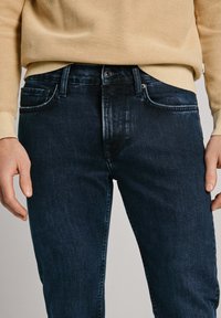 Temno modri traper hlače z oprijetim krojem, ki vključujejo klasičen pet-pocket slog in rahlo zbledel teksturo. Uporabljene z svetlo rumenim puloverjem.