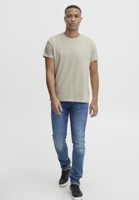 Beige kortermet t-skjorte laget av bomull, kombinert med blå slim-fit jeans og svarte joggesko, båret av en stående mannemodell.