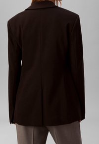 Personne portant un blazer noir cintré et un pantalon gris clair, visible de dos sur un fond gris uni.