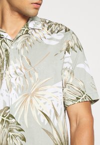 Mies, joka pukeutuu vaaleanvihreään lyhythihaiseen paitaan, jossa on beigen ja valkoisen trooppisen lehden kuvio, näkyy kaulasta puoliväliin rintakehää ja yläkät.