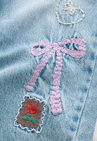 Denim bleu clair avec un nœud brodé violet et un cœur. Comprend un patch avec une fleur rouge sur fond marron, encadré par une couture blanche.