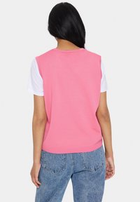 Kvinna med långt svart hår iklädd en rosa ärmlös tröja över en vit t-shirt och blå jeans, vänd bort med en enhetlig bakgrund.