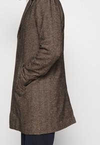 Brauner Mantel mit Fischgrätmuster und tailliertem Schnitt, ausgestattet mit einem angeritzten Reverskragen und Seitentaschen, gefertigt aus strukturiertem Stoff.