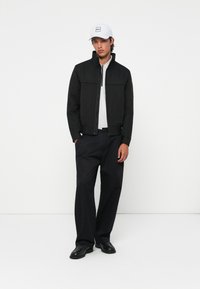 Black jacket with a high collar, gray shirt, black pants, and black shoes. White cap with logo. Simple, modern design.