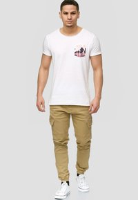 Fehér, rövid ujjú póló, rózsaszín pálmafa grafikával a mellzsebén, bézs cargo nadrággal és fehér sportcipővel párosítva.