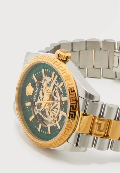 Versace skeletas laikrodis su auksine ir sidabrine metaline apyranke, auksiniu žiedu su graikų rakto raštu ir matomais vidiniais mechaniniais krumpliais.