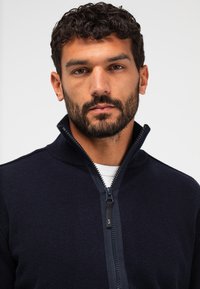 Marineblauer Zip-Up-Pullover mit Stehkragen, ausgestattet mit einem kontrastierenden Reißverschluss und glatter Textur. Über einem weißen Hemd getragen.