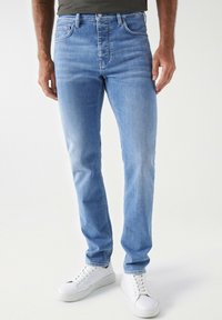Jeans de mezclilla azul claro con corte slim, tiro medio y un ligero desgaste. Diseño de cinco bolsillos con costuras en contraste y cierre de botón. Zapatillas blancas.