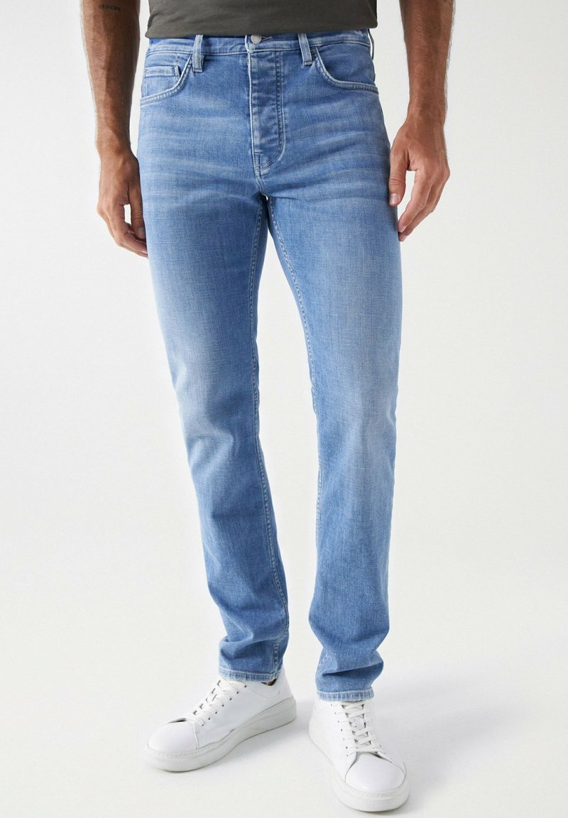 Jeans de mezclilla azul claro con corte slim, tiro medio y un ligero desgaste. Diseño de cinco bolsillos con costuras en contraste y cierre de botón. Zapatillas blancas.