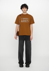 Brown cotton t-shirt with "J'ADORE ARIES" printed in silver letters, paired with black wide-leg jeans and black platform shoes.