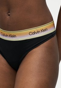 Κοντινό ενός ατόμου που φοράει μαύρα εσώρουχα με ζώνη μέσης σε κλίση από κίτρινο σε μωβ, με επαναλαμβανόμενο κείμενο "Calvin Klein".