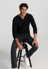 Maglietta henley nera a maniche lunghe con dettagli sui bottoni, abbinata a jeans scuri e sneakers bianche, seduta su uno sgabello nero di fronte a uno sfondo semplice.