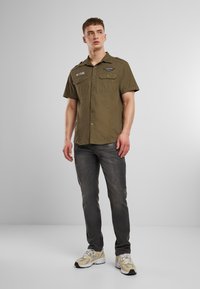 Kortermet olivengrønn skjorte med to brystlommer, knappelukking og skulderklaffer, kombinert med slitne grå jeans og beige joggesko.