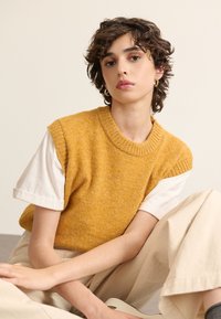 Ein senfgelbes, ärmelloses Strickoberteil mit geripptem Halsauschnitt über einem weißen T-Shirt mit kurzen Ärmeln, kombiniert mit beigefarbenen, schmal zulaufenden Hosen.