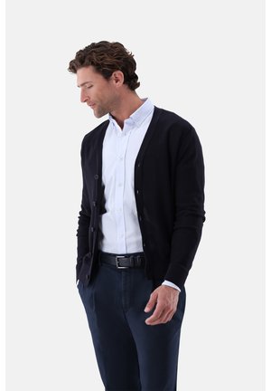 Mann trägt einen schwarzen Cardigan über einem weißen Hemd mit Knöpfen und dunkle Hose, steht mit den Händen in den Taschen und schaut nach unten zu seiner linken Seite.