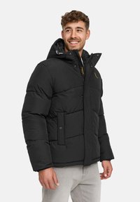 Mand der smiler, iført en sort vinterjakke med hætte og lysegrå bukser, stående mod en ensfarvet lys baggrund.