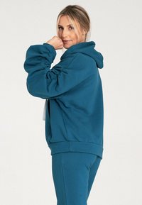 Turkusowa bluza z luźnym krojem, zbieranymi rękawami i kieszenią z przodu, zestawiona z pasującymi spodniami, wykonana z miękkiej mieszanki bawełny.