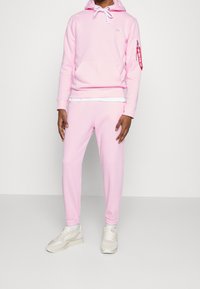 Person i en ljusrosa huvtröja och matchande joggers, vita sneakers, som står mot en enkel ljusgrå bakgrund.