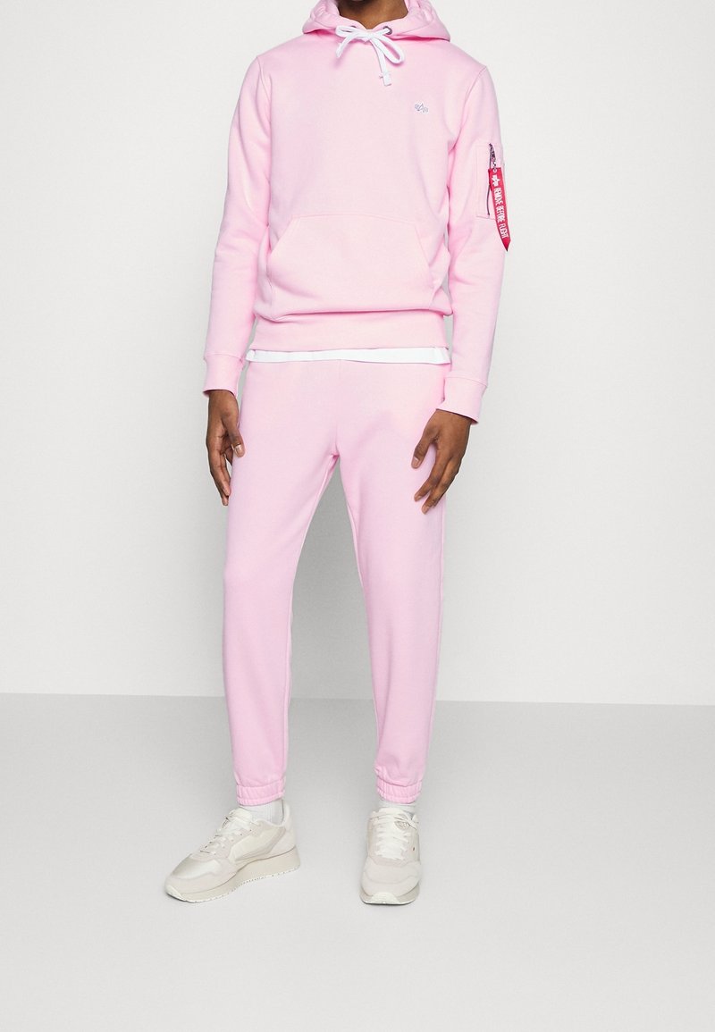 Person i en ljusrosa huvtröja och matchande joggers, vita sneakers, som står mot en enkel ljusgrå bakgrund.