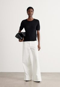 Black knit top with short sleeves and a round neckline, paired with white wide-leg trousers, holding a black pouch bag. Smooth textures.