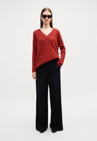 Czerwony sweter ściągaczowy z dekoltem w serek, zestawiony z czarnymi spodniami o szerokich nogawkach i wysokim stanem. Model nosi ciemne okulary przeciwsłoneczne i kolczyki w kształcie kółek.