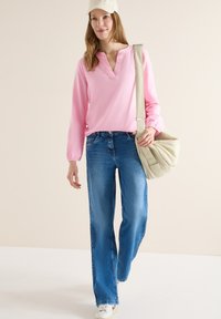 Femme portant un chemisier rose clair, un jean bleu, des baskets blanches et une casquette beige, avec un grand sac à main beige porté à l'épaule.