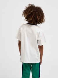 Dziecko o kręconych włosach, ubrane w luźny biały t-shirt i zielone spodnie, stoi tyłem na jednolitym jasnym tle.