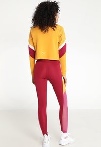 Żółty crop top z siateczkowymi panelami, połączony z wysokimi bordowymi legginsami o kolorowym blokowym wzorze i przezroczystymi akcentami. Białe sneakersy.