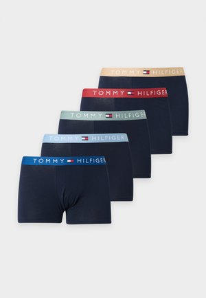 Homme torse nu debout pieds nus portant un boxer foncé avec une ceinture bleu clair sur un fond blanc uni.