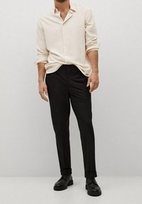 Homme portant une chemise crème à manches longues boutonnée, un pantalon noir sur mesure et des chaussures noires à lacets, debout devant un fond uni clair.