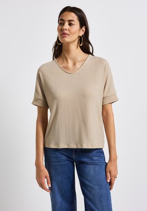 Beige kortærmet top med V-hals, struktureret stof, afslappet pasform og oprullede ærmer, parret med blå denimjeans.