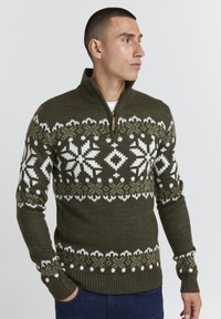 Pull en tricot vert olive avec un col haut et une fermeture éclair, orné de motifs de flocons de neige blancs et vert clair sur la poitrine et les manches.