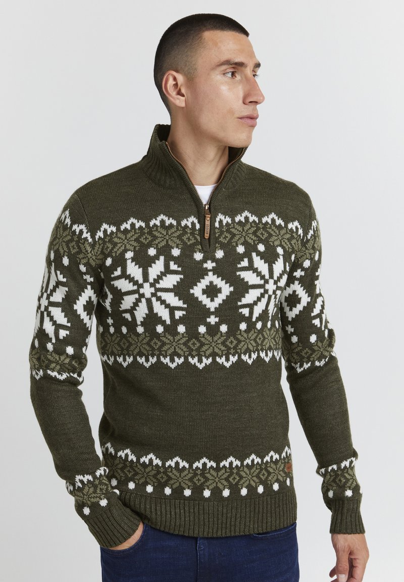 Pull en tricot vert olive avec un col haut et une fermeture éclair, orné de motifs de flocons de neige blancs et vert clair sur la poitrine et les manches.