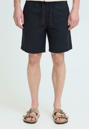 Man wearing black knee-length shorts and beige buckle sandals, standing against a plain light background.
