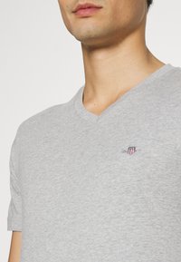 Nahaufnahme einer Person, die ein hellgraues V-Ausschnitt T-Shirt mit einem kleinen bestickten Schild-Logo auf der Brust trägt.