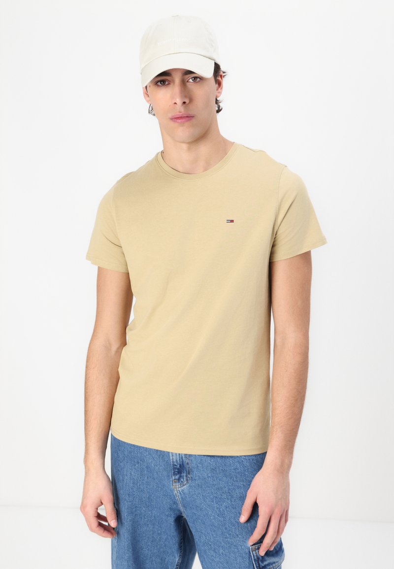 Camiseta de algodón beige con mangas cortas y escote redondo, que presenta un pequeño logotipo bordado. Llevada con jeans de mezclilla azul claro.