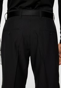 Czarne spodnie o gładkiej fakturze, z dwiema tylnymi kieszeniami zapinanymi na guziki oraz eleganckim, dopasowanym kroju. W zestawie znajduje się pasujący czarny pasek.