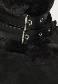 Zwarte nepbont kraag met een leren band, voorzien van twee zilveren gespen en oogjes, wat zorgt voor een textuurcontrast en een veilige pasvorm.